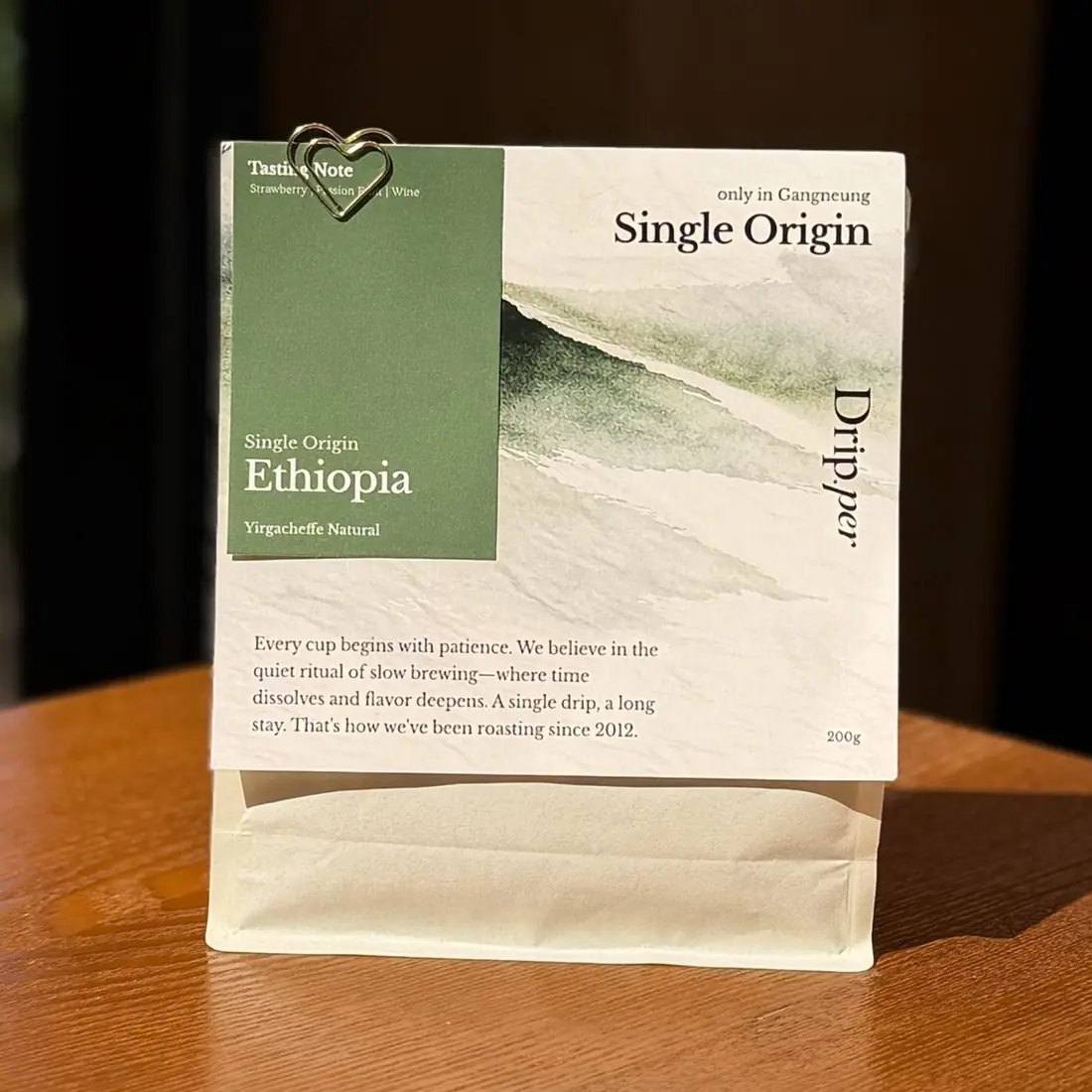 우드 테이블에 세워 둔 Drip.per 싱글 오리진 에티오피아 200g 원두 봉투. 진한 초록 택에 Ethiopia, Yirgacheffe Natural 이라고 적혀 있고 향미 노트는 Strawberry, Passion Fruit, Wine 이다.