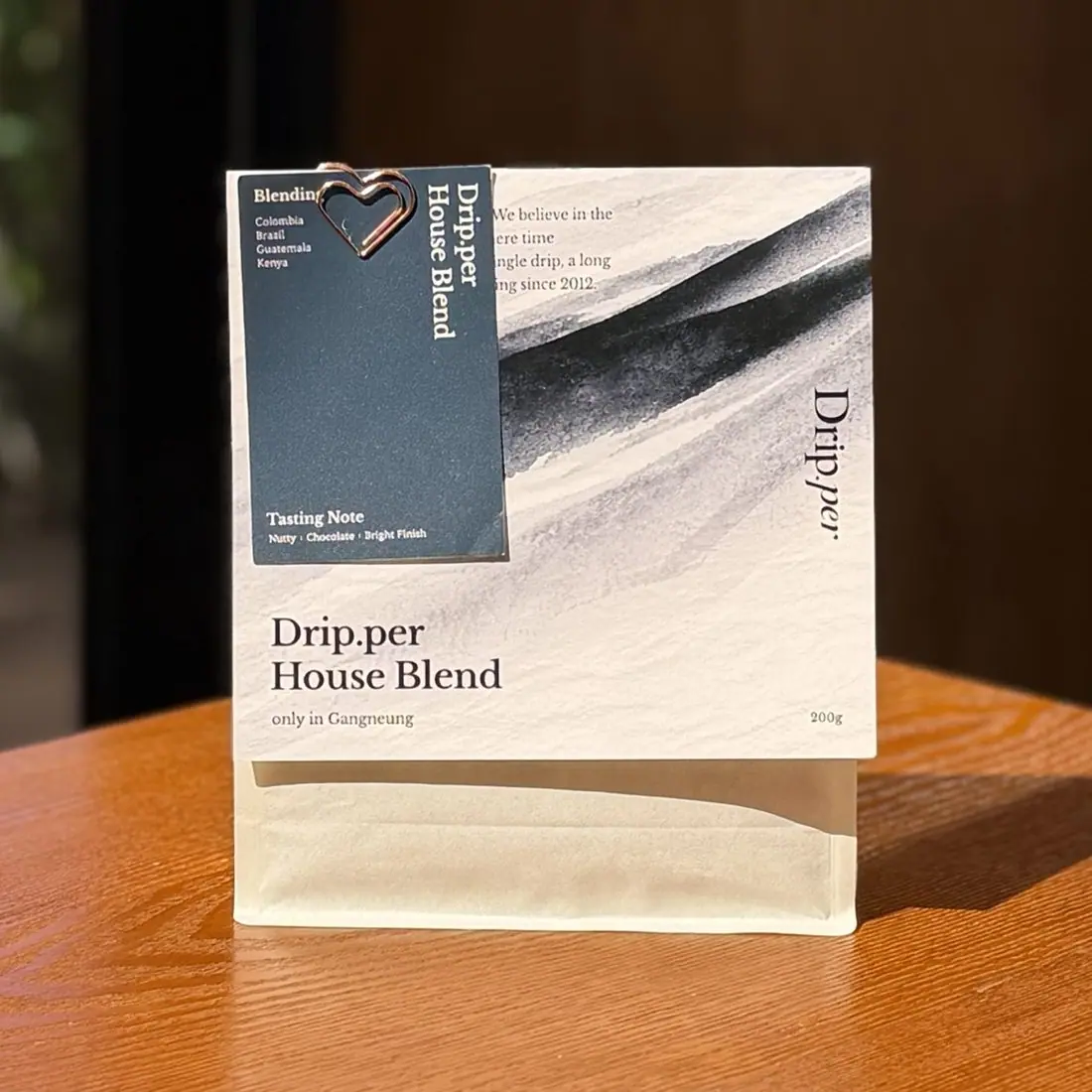우드 테이블에 세워 둔 Drip.per House Blend 200g 원두 봉투. 남색 택에 블렌딩 산지로 Colombia, Brazil, Guatemala, Kenya 가 적혀 있고 향미 노트는 Nutty, Chocolate, Bright Finish 다.