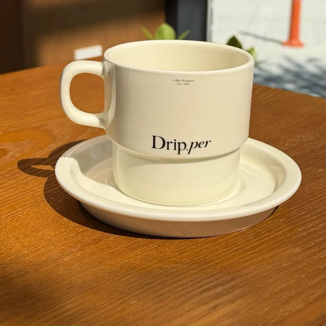 우드 테이블에 놓인 Drip.per 로고가 새겨진 크림색 도자기 잔과 받침 접시.